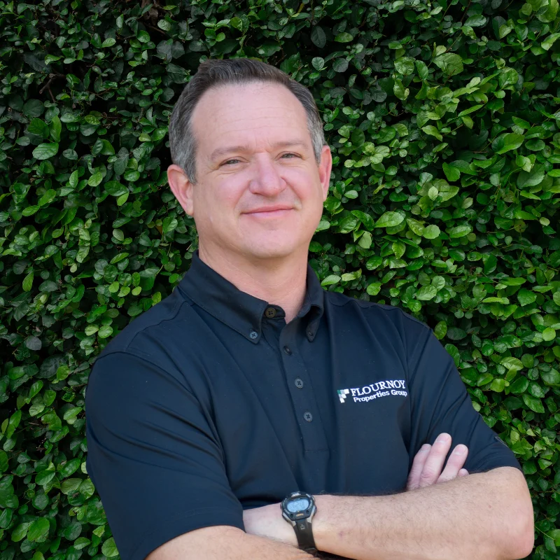 A man with short hair smiles confidently, wearing a black polo shirt against a lush green background.