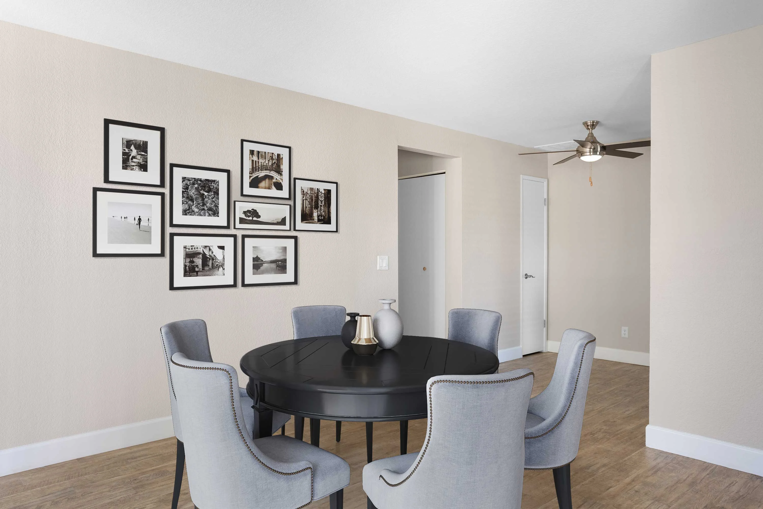 A modern dining room featuring a round black table surrounded by six light gray chairs. The walls are painted a soft beige and decorated with a gallery of black and white framed photographs. A ceiling fan is visible on the ceiling, and there is a doorway leading to another room.