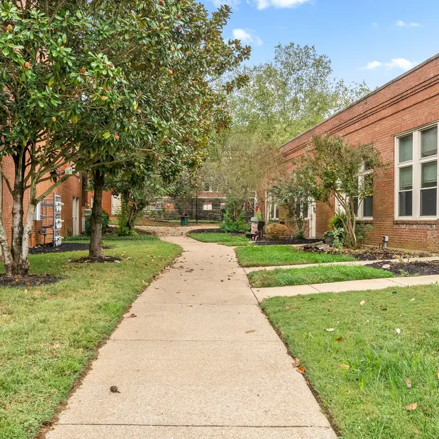 A calm residential pathway flanked by brick buildings and greenery, with a clear blue sky overhead.