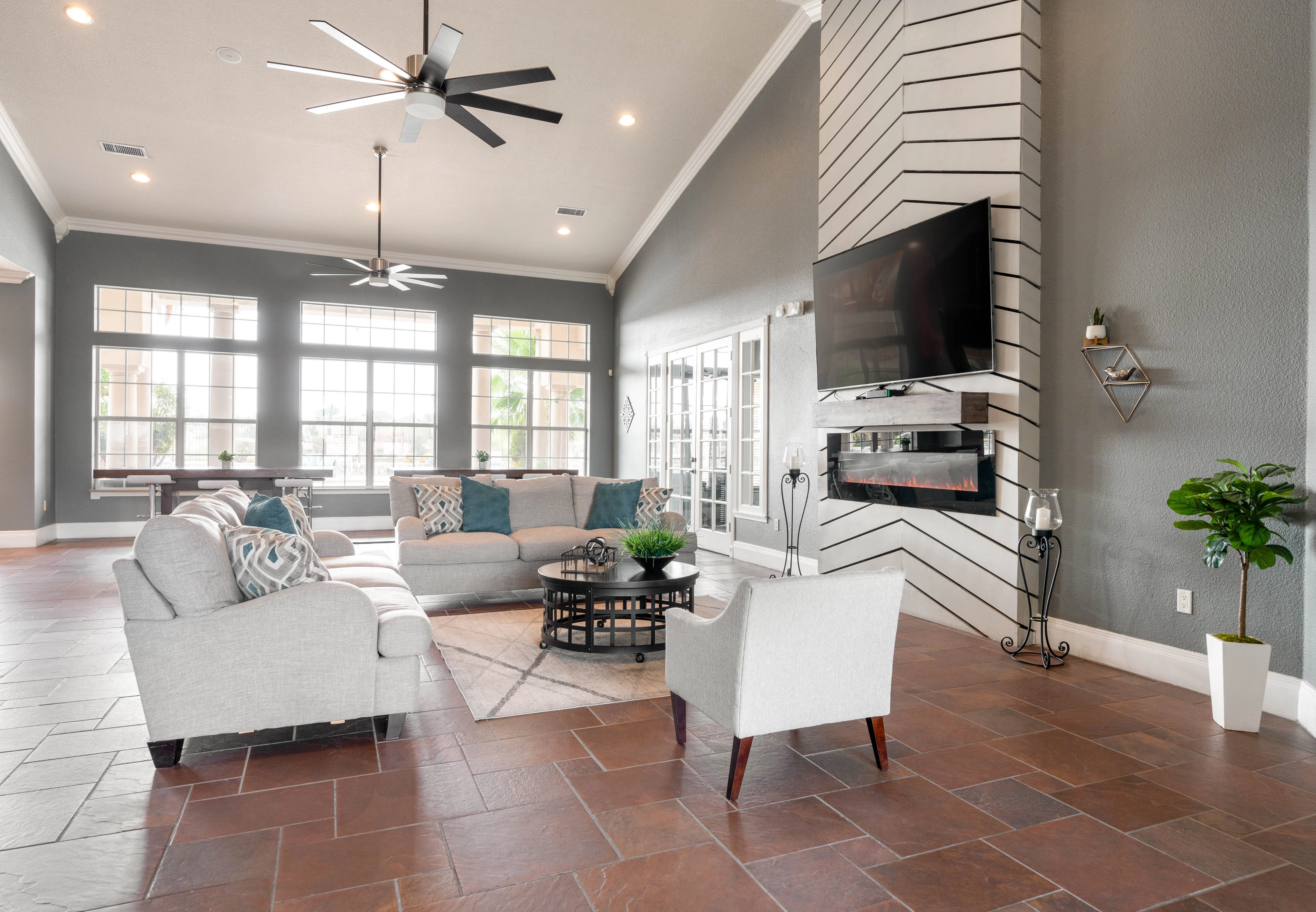 A spacious community living area featuring a large sectional sofa, a coffee table, and a modern fireplace. There are large windows allowing natural light to flood the space, alongside ceiling fans and decorative plants.