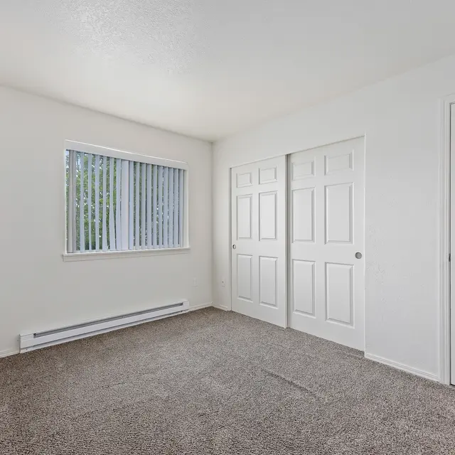 Vacant Room Interior A vacant room featuring beige carpet flooring, white walls, and a window with vertical blinds. There are two closed double doors on one side of the room, and a single door on the other. The room is well-lit with natural light.