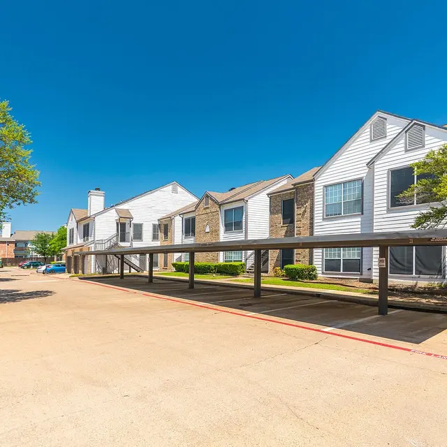 An exterior view of an apartment complex with two-tone buildings and carports under a clear blue sky. Trees are visible on the left side, and a driveway runs through the scene.