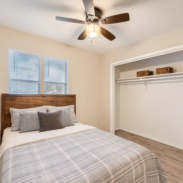 A cozy bedroom featuring a bed with a plaid blanket, a wooden headboard, a nightstand, and a tall green plant. Natural light comes through two windows, and a closet is seen in the background with open shelves and baskets.
