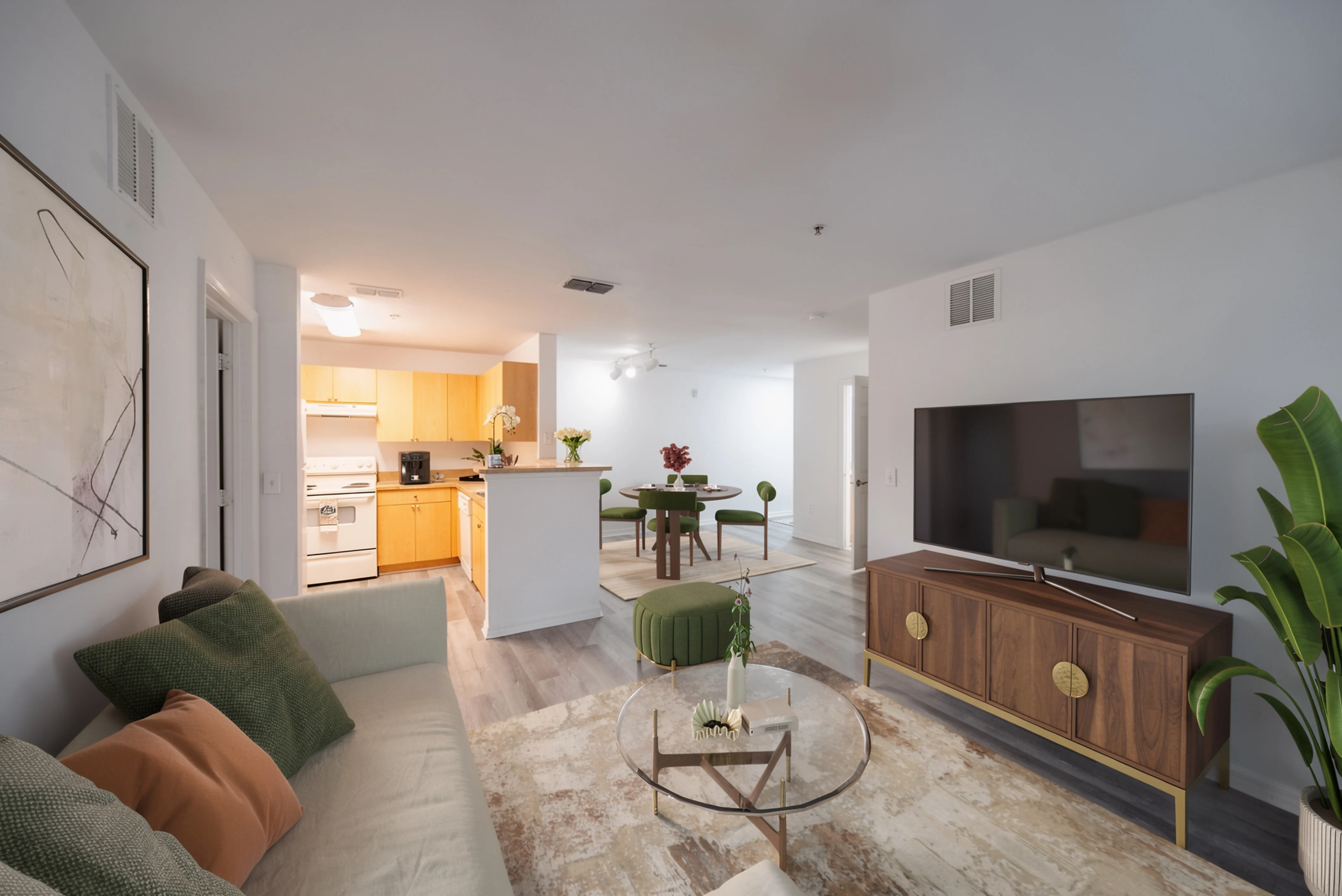 A spacious modern apartment interior featuring a living room with a light sofa, coffee table, and a television mounted on the wall. The open kitchen is visible in the background, showcasing wooden cabinetry and white appliances. A dining area with green chairs is also present, along with decorative elements like plants and a flower arrangement.