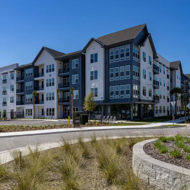 A modern apartment building with a mix of white and dark gray exterior, surrounded by landscaping and a parking area. The sky is clear and blue, indicating a sunny day.