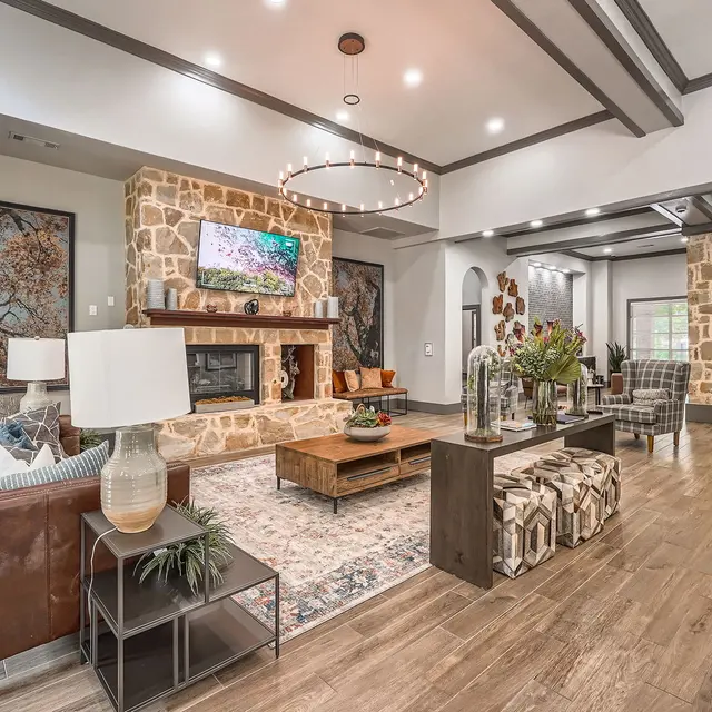 A modern living room featuring a stone fireplace, large TV, and stylish furniture. The space has wooden flooring and a neutral color palette, complemented by artwork and decorative plants.