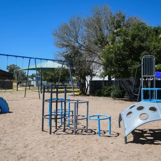 A playground featuring a swing set, a slide, and climbing equipment on sandy ground, surrounded by trees and a building.