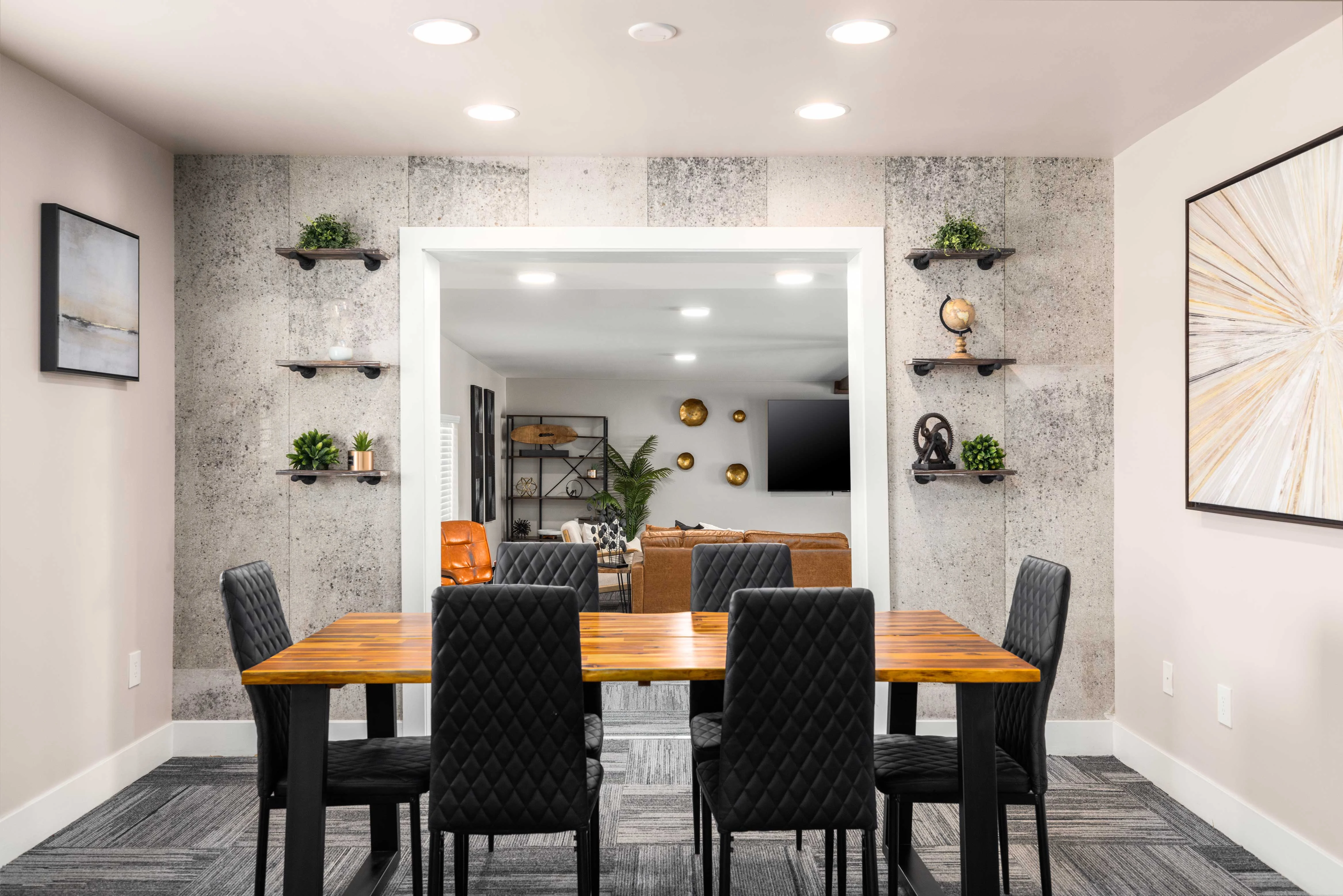 A modern dining area with a wooden table surrounded by black quilted chairs. The walls are decorated with gray concrete panels and decorative shelves with potted plants. A doorway opens to a living space with a seating arrangement and a TV.