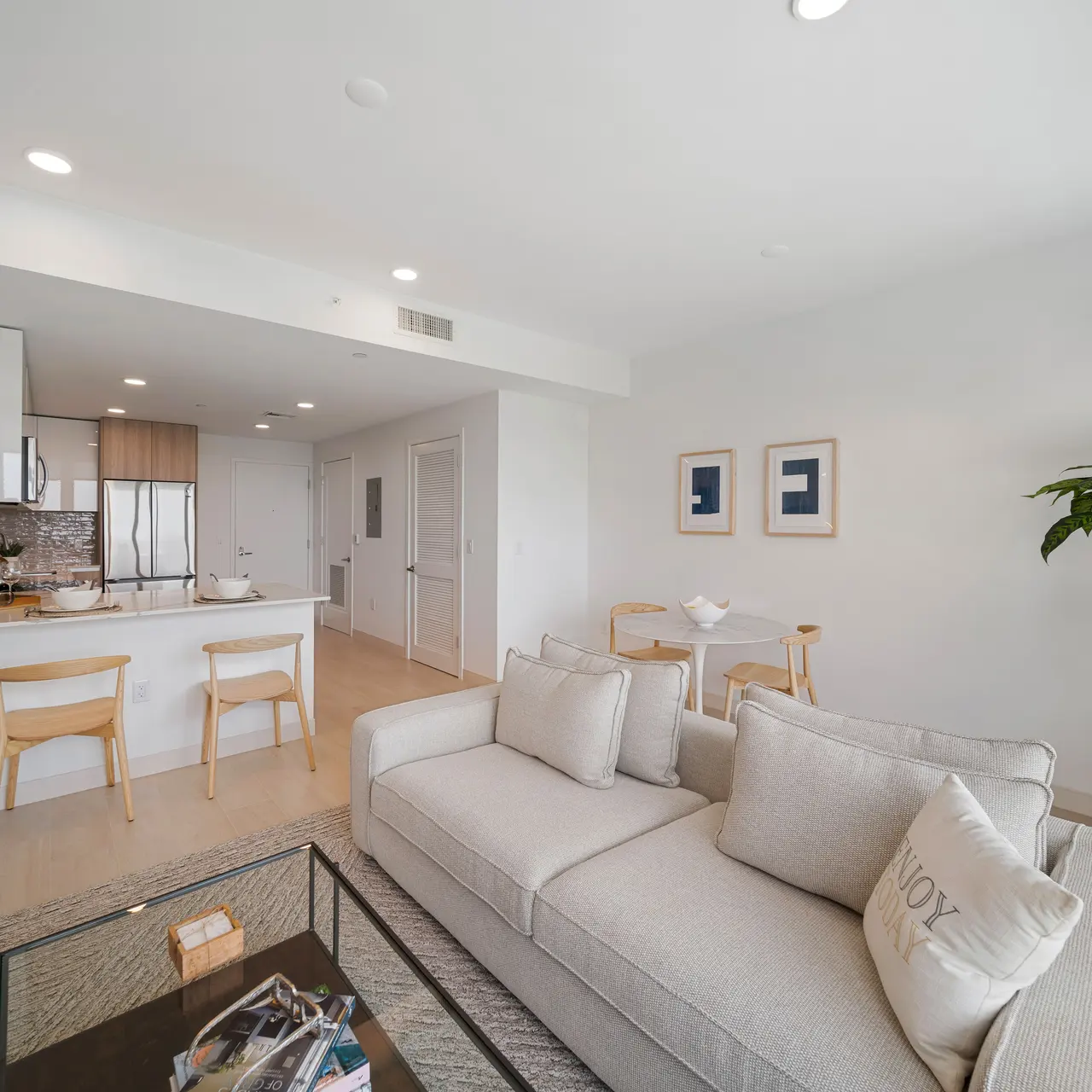 A modern apartment living room featuring a light sofa, glass coffee table, and a potted plant. The kitchen area is visible in the background with sleek cabinetry and bar stools.