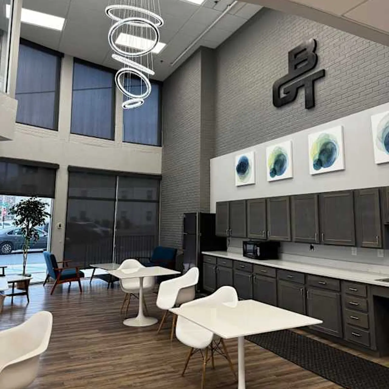 A stylish modern office interior featuring a kitchen area, white tables, and chairs, with artistic wall decor and large windows allowing natural light.