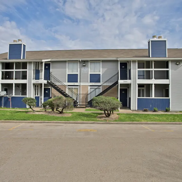 A two-story apartment complex featuring multiple units with blue and gray exterior, small shrubs in front, and a pathway leading up to the entrance.