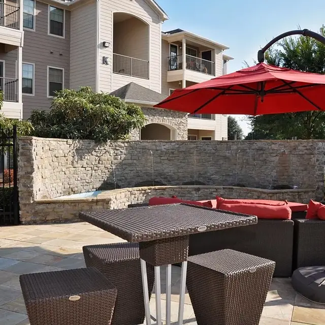 An outdoor lounge area with a red umbrella over a table and chairs, surrounded by greenery and a stone wall, with apartments in the background.