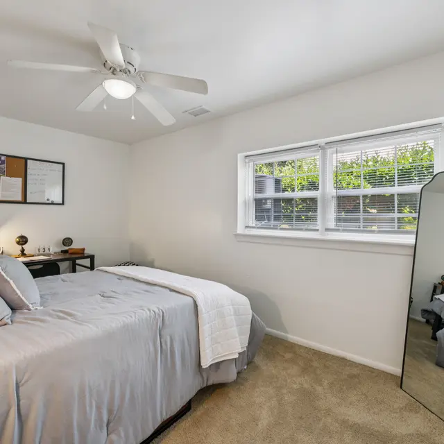 Cozy Bedroom Interior A cozy bedroom featuring a bed with light gray bedding, a ceiling fan, a mirror, and windows with greenery outside.