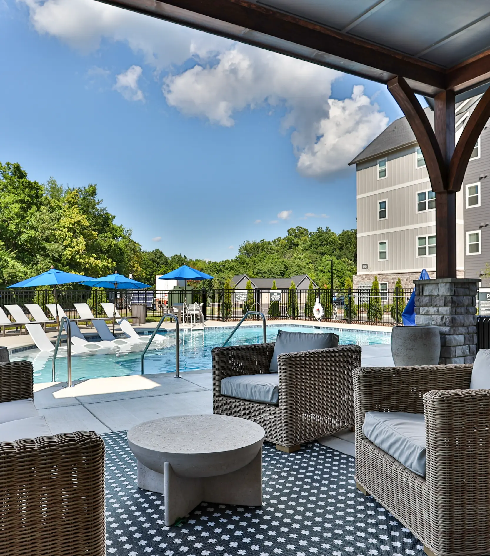 A pool area featuring chairs and shaded umbrellas, surrounded by greenery under a blue sky with fluffy clouds.