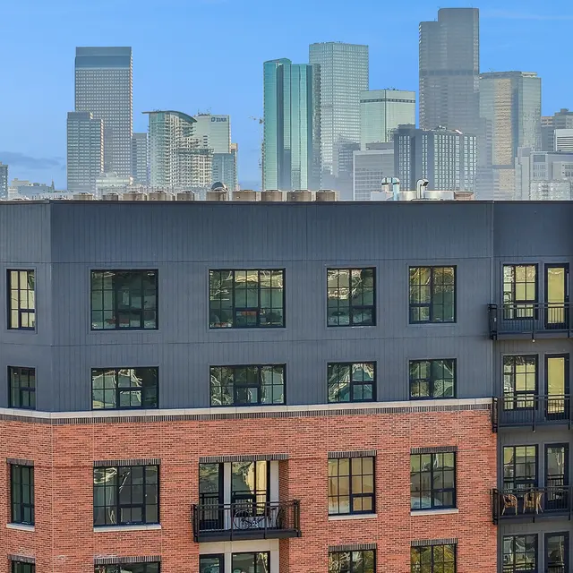 Urban Apartment Living A view of modern apartment buildings in the foreground with a city skyline in the background. The building features a mix of red brick and dark exterior, with balconies visible. A leasing sign is prominently displayed on the building.