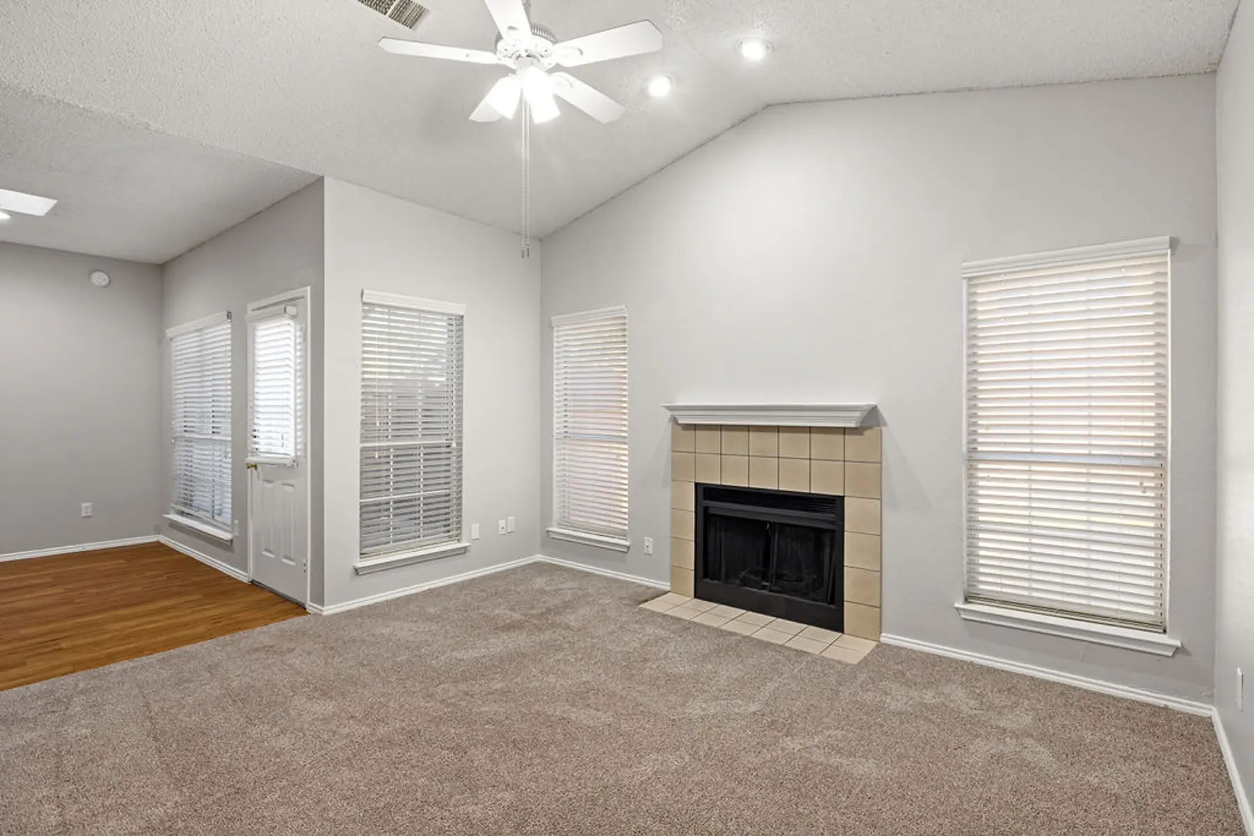 Cozy Living Room Interior A spacious living room featuring light-colored walls, a carpeted floor, and a fireplace. Large windows are adorned with blinds, and there is a ceiling fan overhead. An open area can be seen leading to an entrance with additional windows.