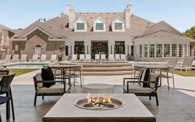 A modern pool area with outdoor seating, featuring a fire pit in the foreground and a large, elegant building in the background.