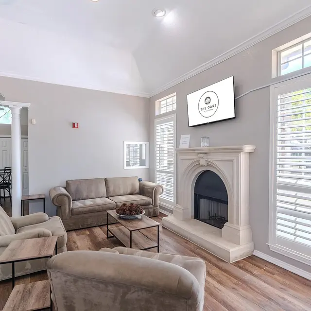 A bright and cozy living room featuring a beige sofa, two matching armchairs, a coffee table, and a fireplace. A flat-screen TV is mounted on the wall, and sunlight streams through the window with white shutters.