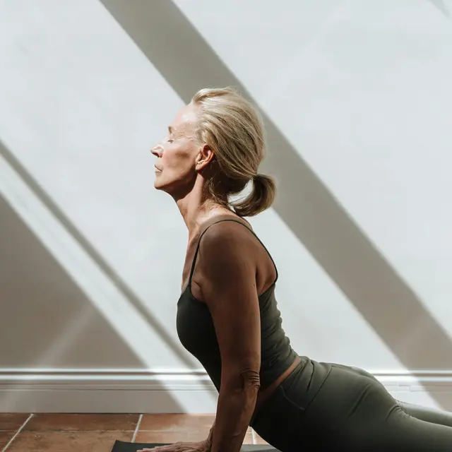 A woman in a yoga pose on a mat, with sunlight casting shadows on the wall behind her.