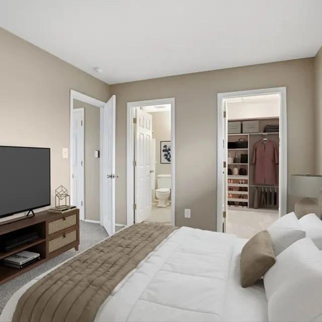 A modern bedroom featuring a large bed with white bedding and a decorative brown throw. A wooden TV stand with a flat-screen television is positioned in front, and there's a wardrobe visible with a closet in the background. The walls are painted a light taupe color, and there is natural light coming through the window.