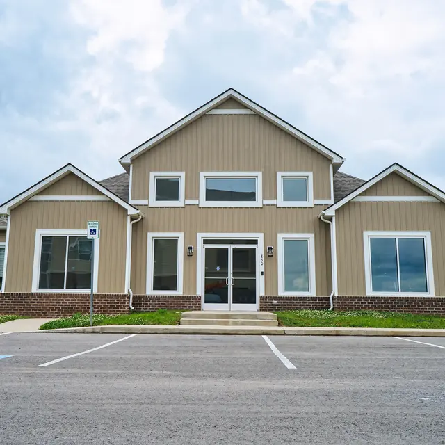 A modern two-story building with a beige exterior, large windows, and a central entrance. The front yard is landscaped with green grass and there are parking spaces visible in front.