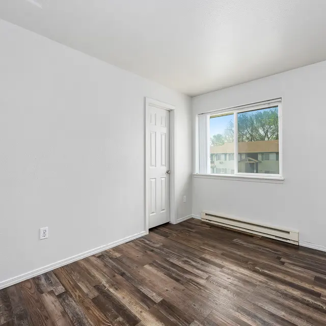 Empty Room Interior A clean, empty room with light-colored walls and dark wooden flooring. There is a window with a view of the outside and a closed door on one wall.
