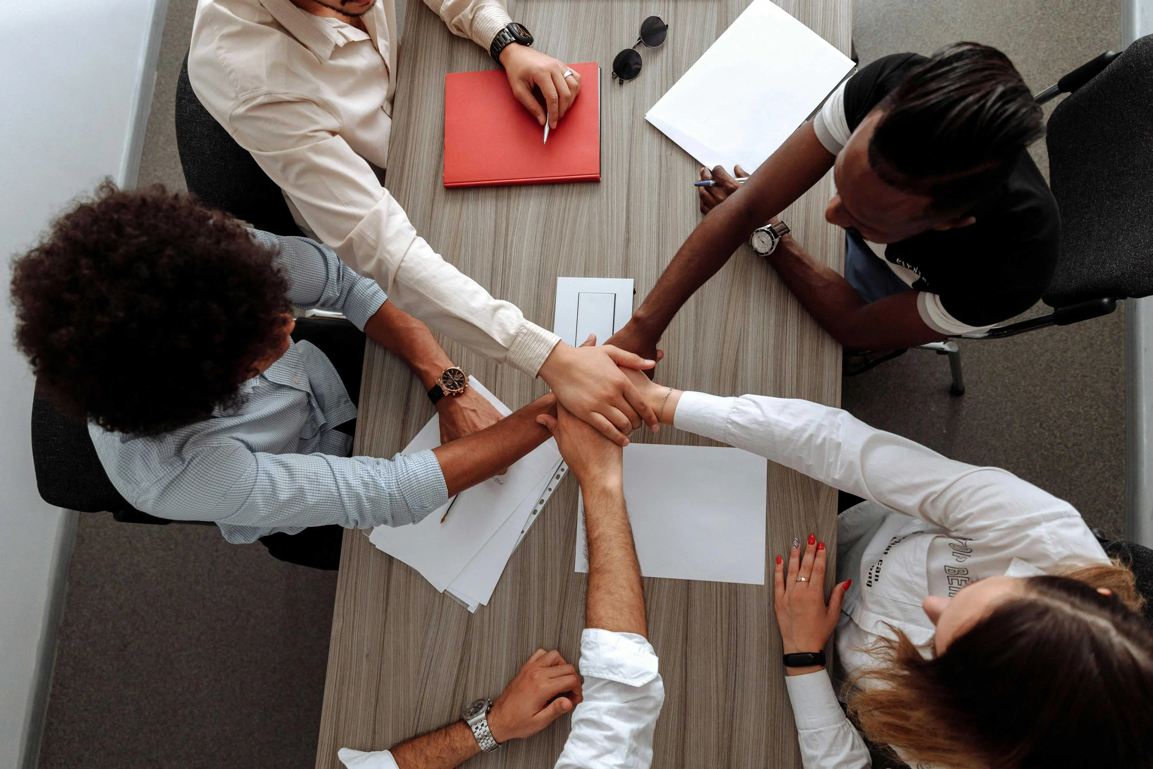 A group of diverse individuals in business attire are placing their hands together on a table in a gesture of teamwork and collaboration. Papers and a red notebook are visible on the table.