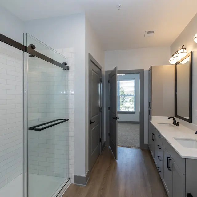 A modern bathroom featuring a glass shower, double sinks, and large mirrors. The decor includes grey cabinetry and a wooden floor.