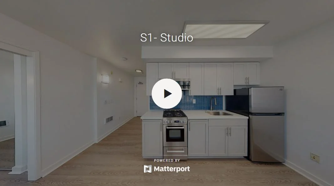 Interior view of a modern studio apartment featuring a kitchen area with stainless steel appliances, white cabinetry, and wood flooring.