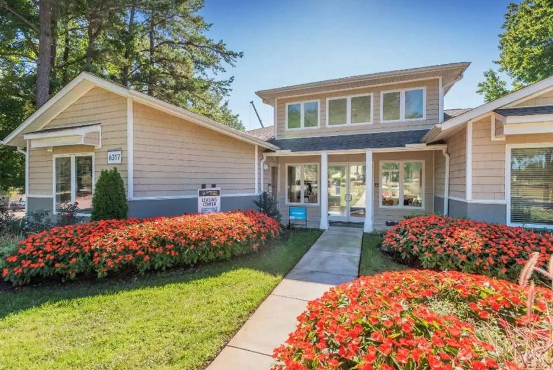 A charming home exterior featuring a light-colored facade with a landscaped pathway. Brightly colored flower beds in the foreground and a clear blue sky in the background.