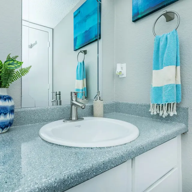 A modern bathroom sink area featuring a white circular sink, a silver faucet, and a light blue and white striped towel hanging on a towel rack. There is a decorative blue and white vase with a green plant, and the countertop is a speckled light gray. In the background, there is a mirror reflecting the space and a door visible.