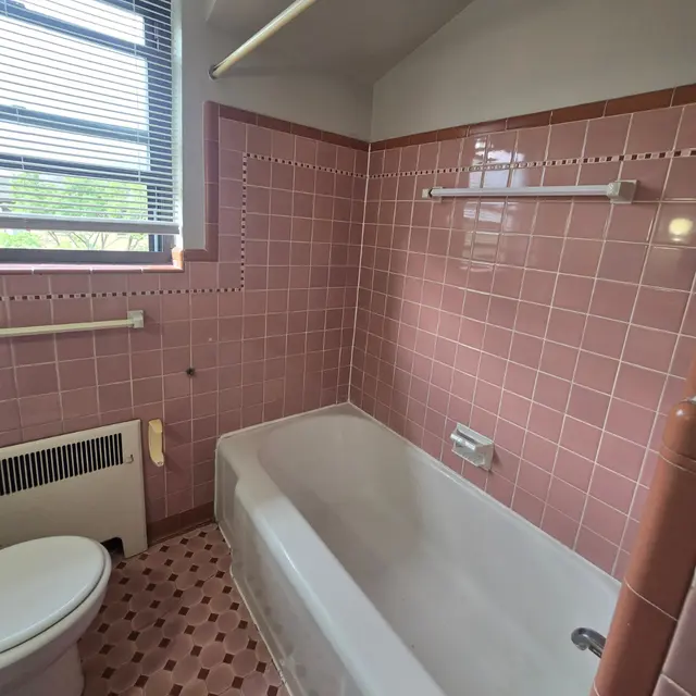 Vintage Pink Bathroom A bathroom with pink tiled walls and floor, featuring a bathtub, a toilet, and a window with blinds.