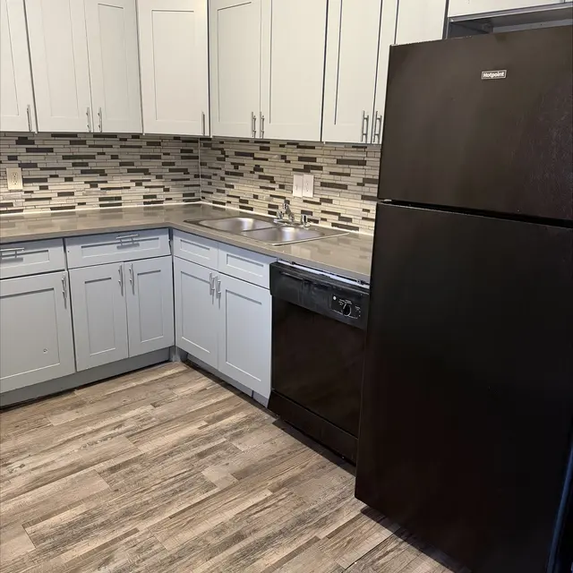 An interior view of a modern kitchen featuring light gray cabinets, a dark black refrigerator, a dishwasher, and a tiled backsplash with a mix of gray and white tones.
