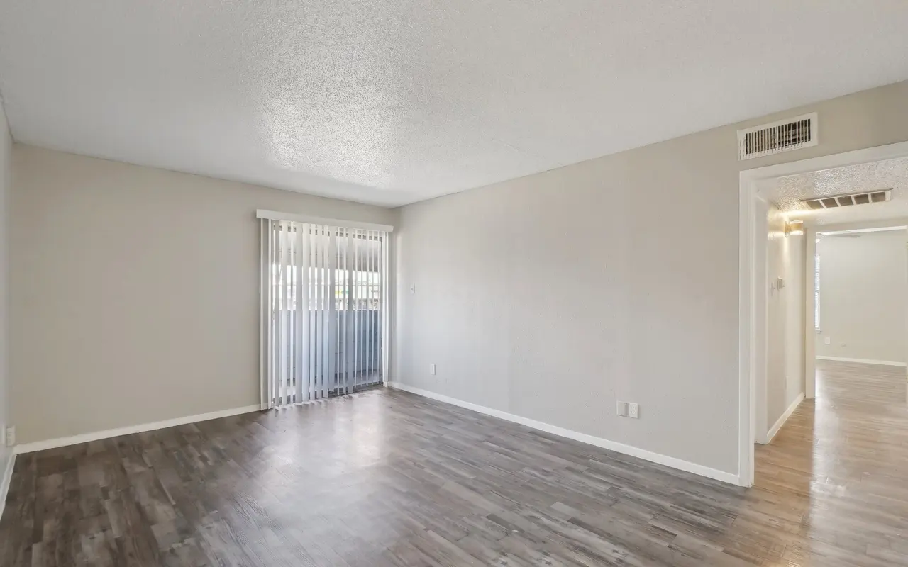 A spacious and empty living room with light gray walls and laminate flooring. There are vertical blinds covering a window, and a doorway leading to another room is visible on the right.