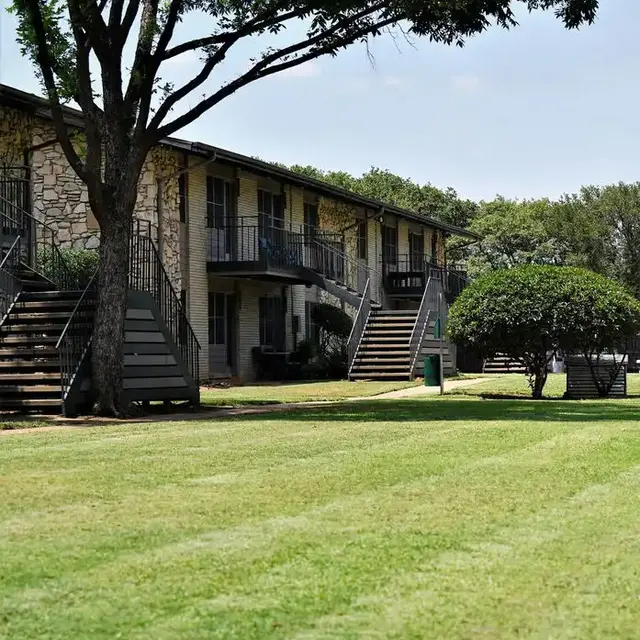 View of a two-story apartment building with exterior staircase, surrounded by green grass and trees.