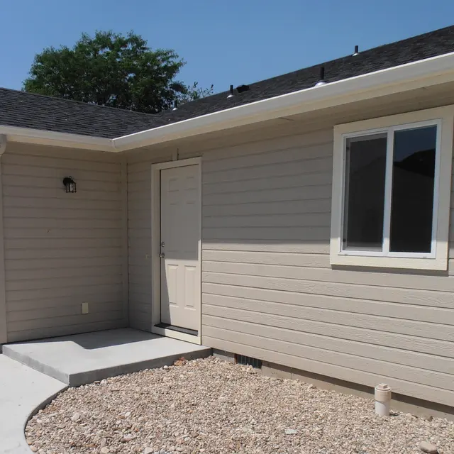 Exterior view of a modern single-story house with a light gray exterior, including a front porch and a door, framed by a gravel yard.