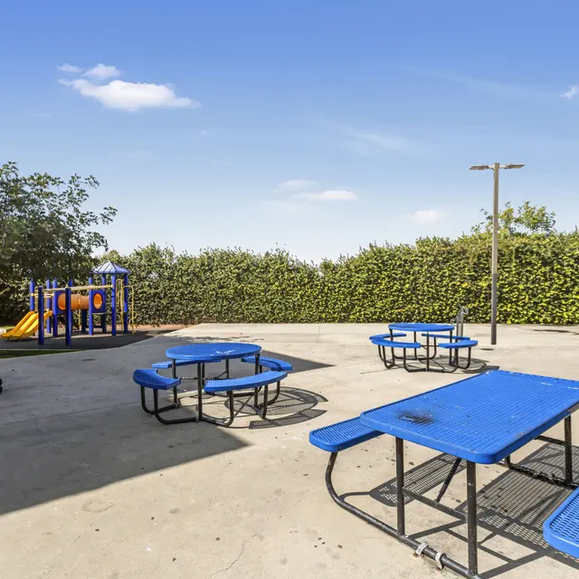 An outdoor playground area with blue picnic tables and a play structure in the background.