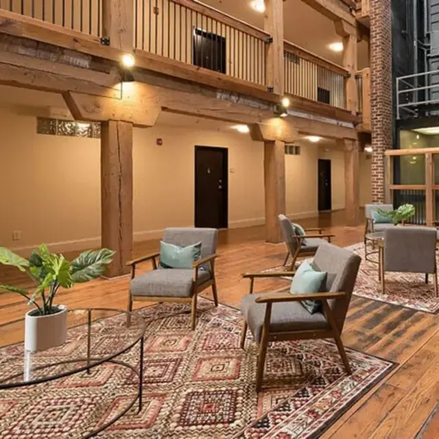 A spacious lobby with wooden floors, featuring comfortable seating in gray chairs, a glass coffee table, and a decorative plant. The walls include warm tones and brick elements, with open areas leading to doors and an elevator in the background.