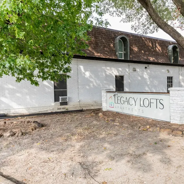 Exterior view of the Legacy Lofts Apartments building featuring a white brick wall sign with the name, surrounded by trees and a rocky ground area.