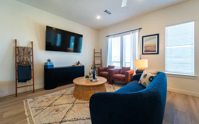 A cozy modern living room featuring a blue sofa, a round coffee table, and two brown armchairs. There's a television mounted on the wall and natural light coming through the windows. A decorative ladder and a rug complete the space.