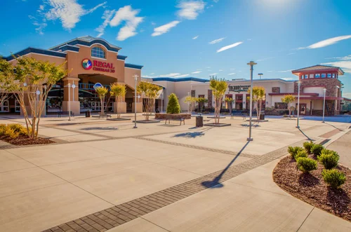 A bright, sunny day at a shopping center featuring a Regal Cinemas building. The plaza has smooth concrete pathways, palm trees, and neatly landscaped areas. Several storefronts are visible in the background under a clear blue sky.