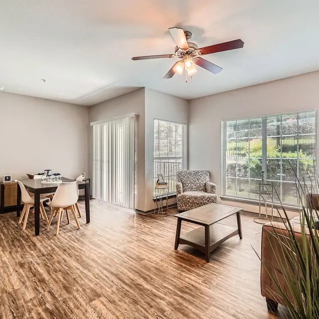 A spacious living room featuring a wooden floor, a ceiling fan, a cozy seating area, and a dining table with chairs. Large windows allow natural light to fill the space, and there's a small balcony.