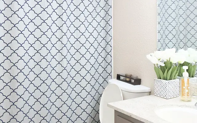 A modern bathroom featuring a white and navy patterned shower curtain, a toilet, and a sink with a beige cabinet. Fresh flowers are placed on the counter beside soap dispensers.