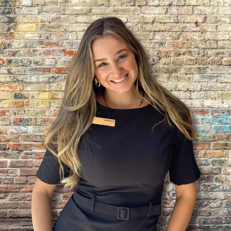 Smiling Receptionist A smiling woman in a black dress stands in front of a rustic brick wall, wearing a name tag that reads 'Ashley'.