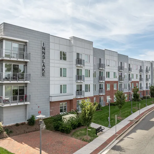 A modern apartment building with multiple stories, featuring a blend of brick and gray siding. The front has balconies and large windows, surrounded by green lawns and landscaped areas. A curved road leads to the building's entrance.
