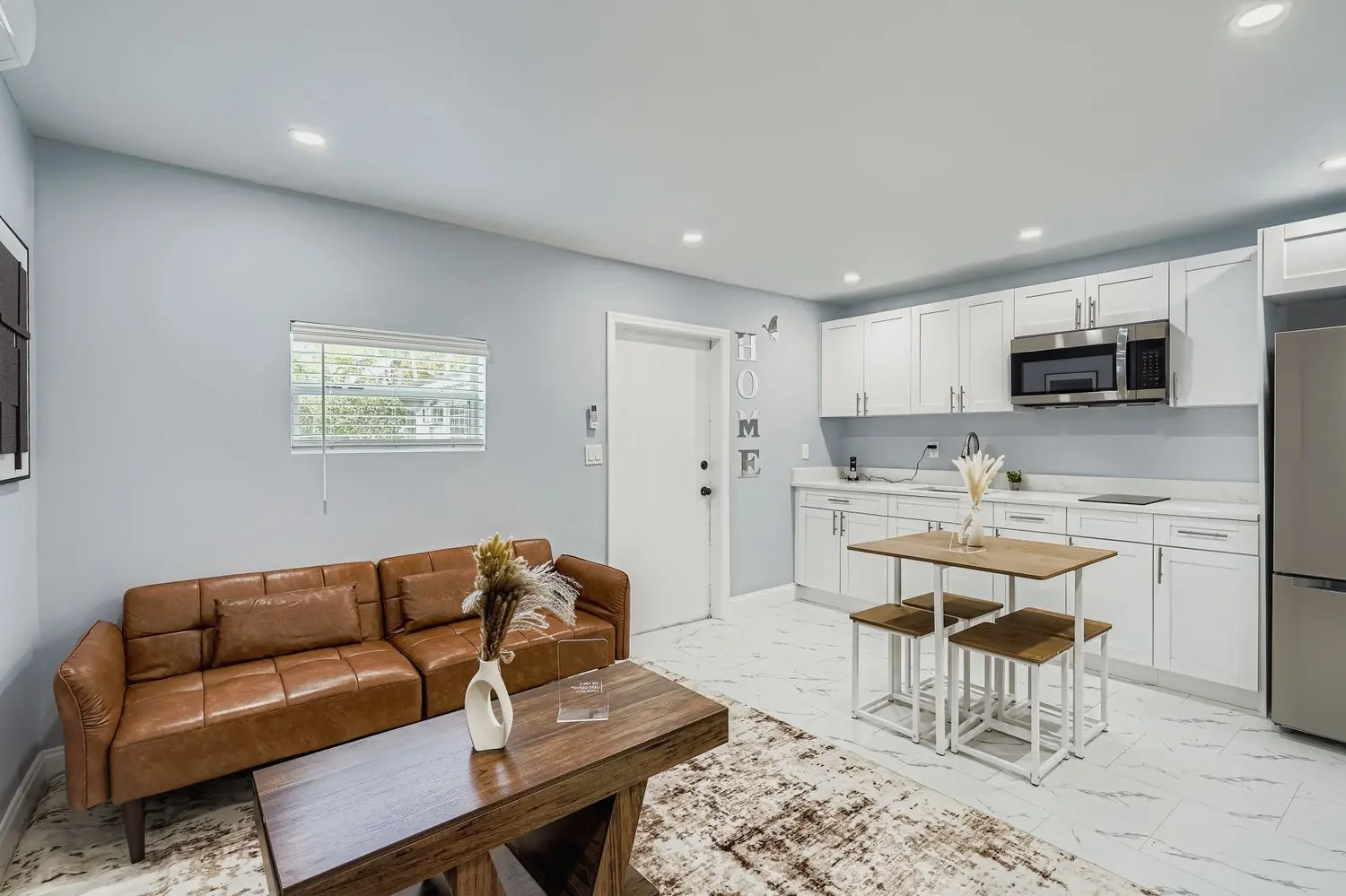 A modern living space featuring a brown leather sofa, a wooden coffee table, and a dining area with four stools. The kitchen is in the background with white cabinets and appliances.