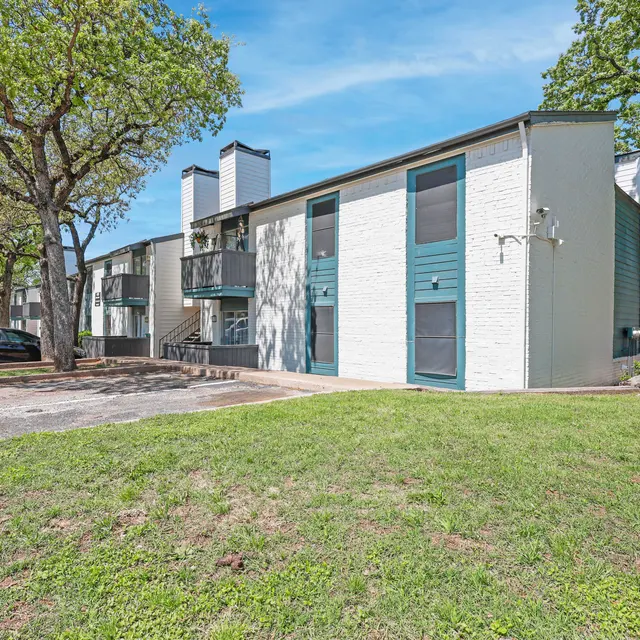Exterior view of a modern, two-story apartment building with a green lawn and trees. The building features blue windows and balconies, with a parking area visible in the foreground.