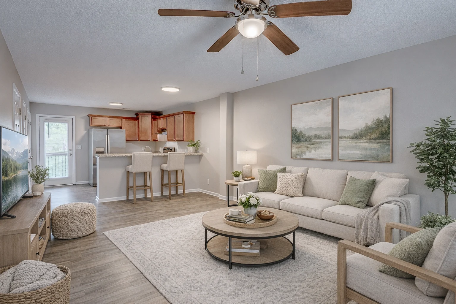 A spacious and inviting modern living room featuring light-colored walls, a comfortable sofa with decorative pillows, a round coffee table, and a cozy area rug. There is a ceiling fan, a flat-screen TV on a media console, and an open kitchen area with bar stools in the background.