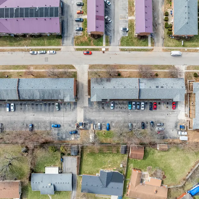Aerial view of an apartment complex featuring multiple low-rise buildings with a large parking lot in the center. The surrounding area includes residential homes and green spaces.