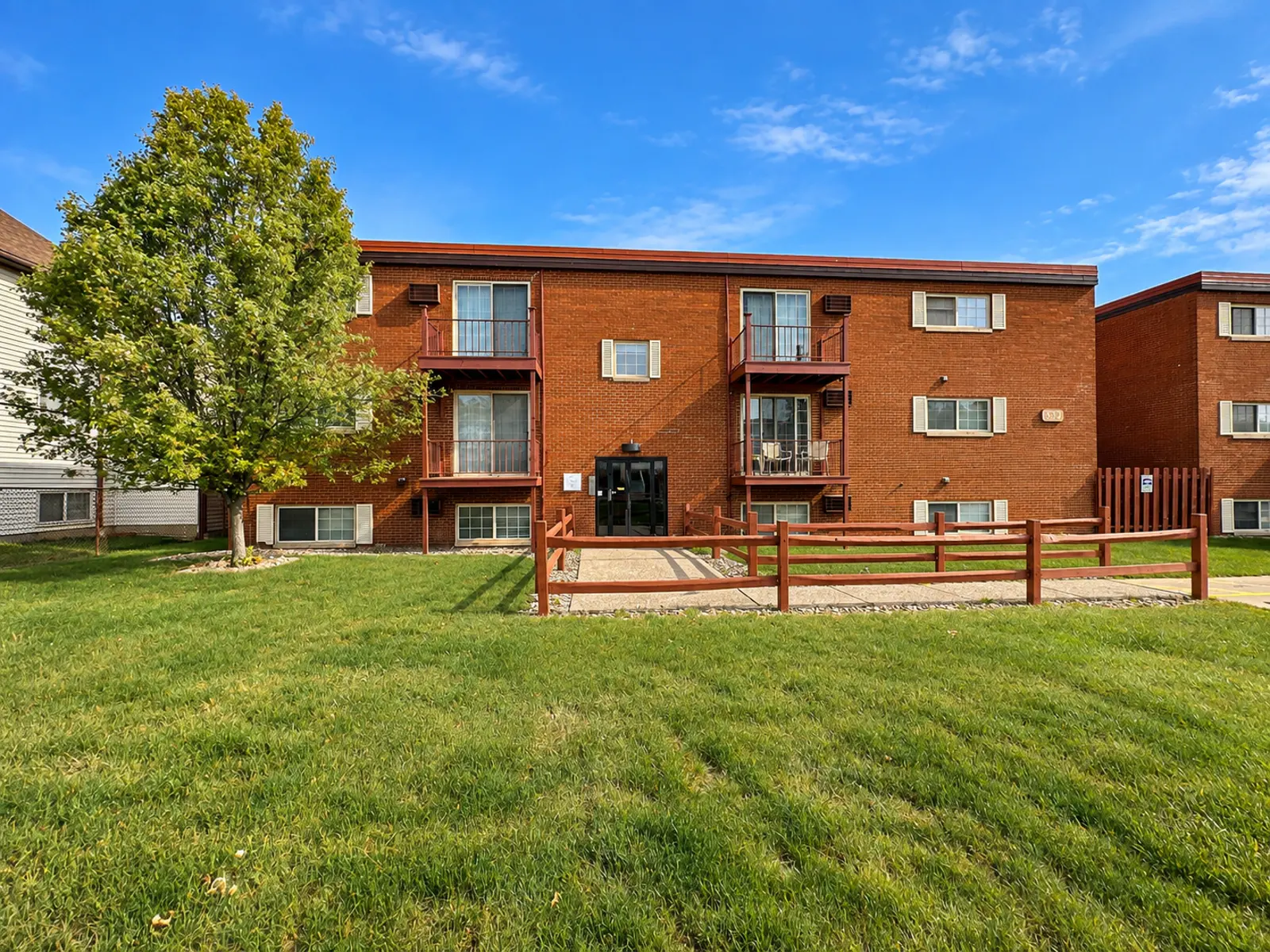 Exterior view of a brick apartment building with balconies surrounded by green grass and trees.